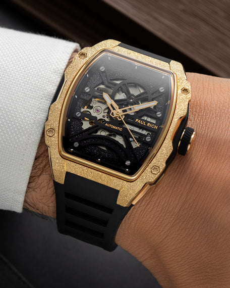 Gold frosted tonneau skeleton watch with black strap, worn on wrist beside a white textured shirt cuff.
