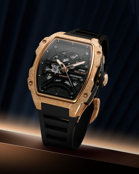 Rose gold frosted tonneau case watch with open skeleton dial and black strap on a dark blue draped background.