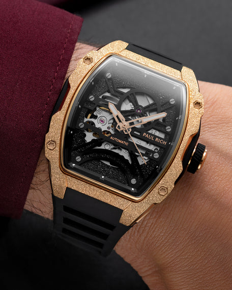 Rose gold frosted tonneau skeleton watch with black strap, shown on wrist beside a burgundy sleeve.