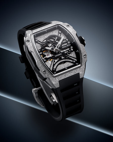Silver frosted tonneau case watch with open skeleton dial and black strap on a dark blue geometric background.