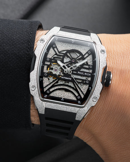 Silver frosted tonneau skeleton watch with black strap, worn on wrist beside a black sleeve.