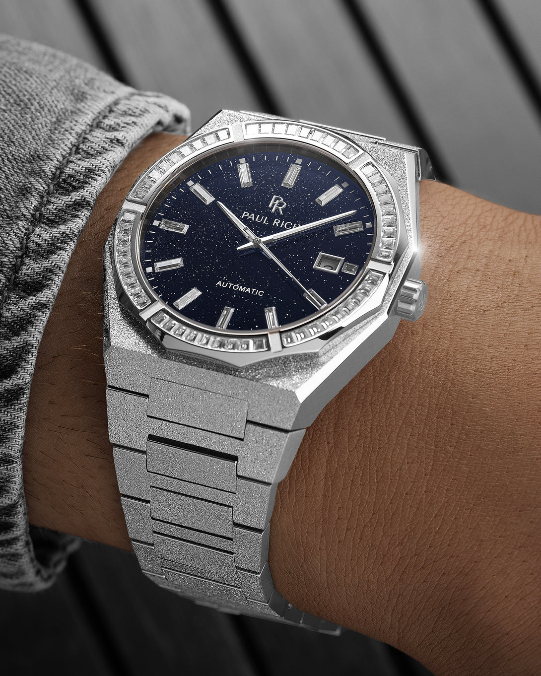 Silver frosted watch with a dark blue aventurine dial and baguette-cut crystal bezel on a wrist wearing a gray textured sleeve.