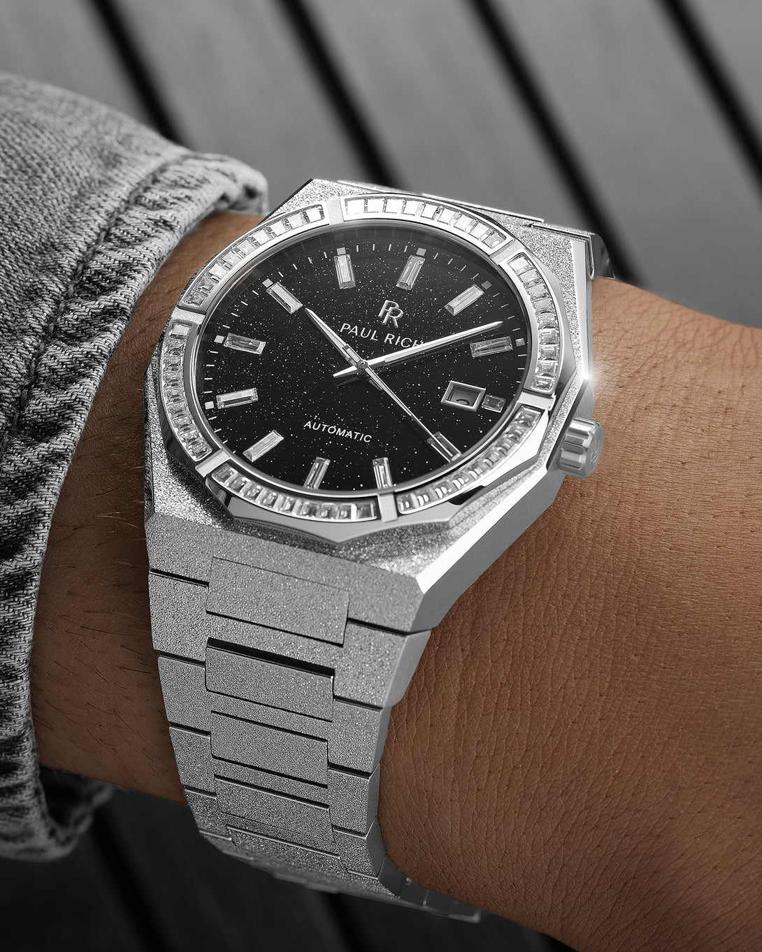 Silver frosted watch on a wrist with a black aventurine dial, baguette-cut stones around the bezel, and a textured silver bracelet.