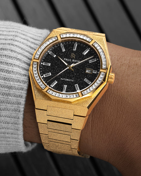 Gold-tone frosted watch on a wrist with a black aventurine dial, baguette-cut stones on the bezel, gold markers, and a textured gold bracelet.