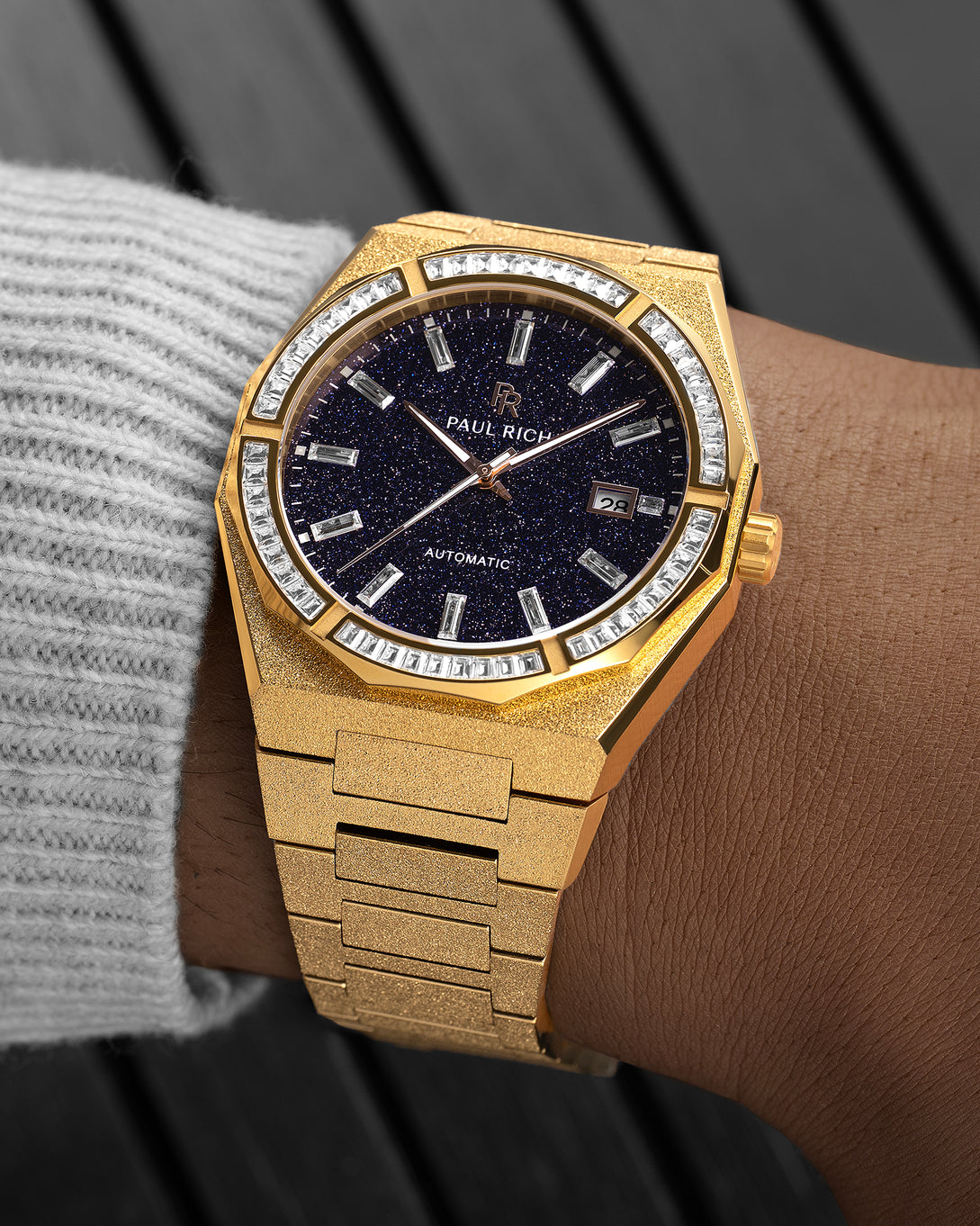 Gold frosted watch with a blue aventurine dial and baguette-cut crystal bezel on a wrist wearing a light gray knit sleeve.