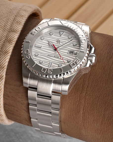 Stainless steel watch with a white wave-textured dial and red seconds hand shown on a wrist, highlighting the bezel, bracelet, and brushed finish.