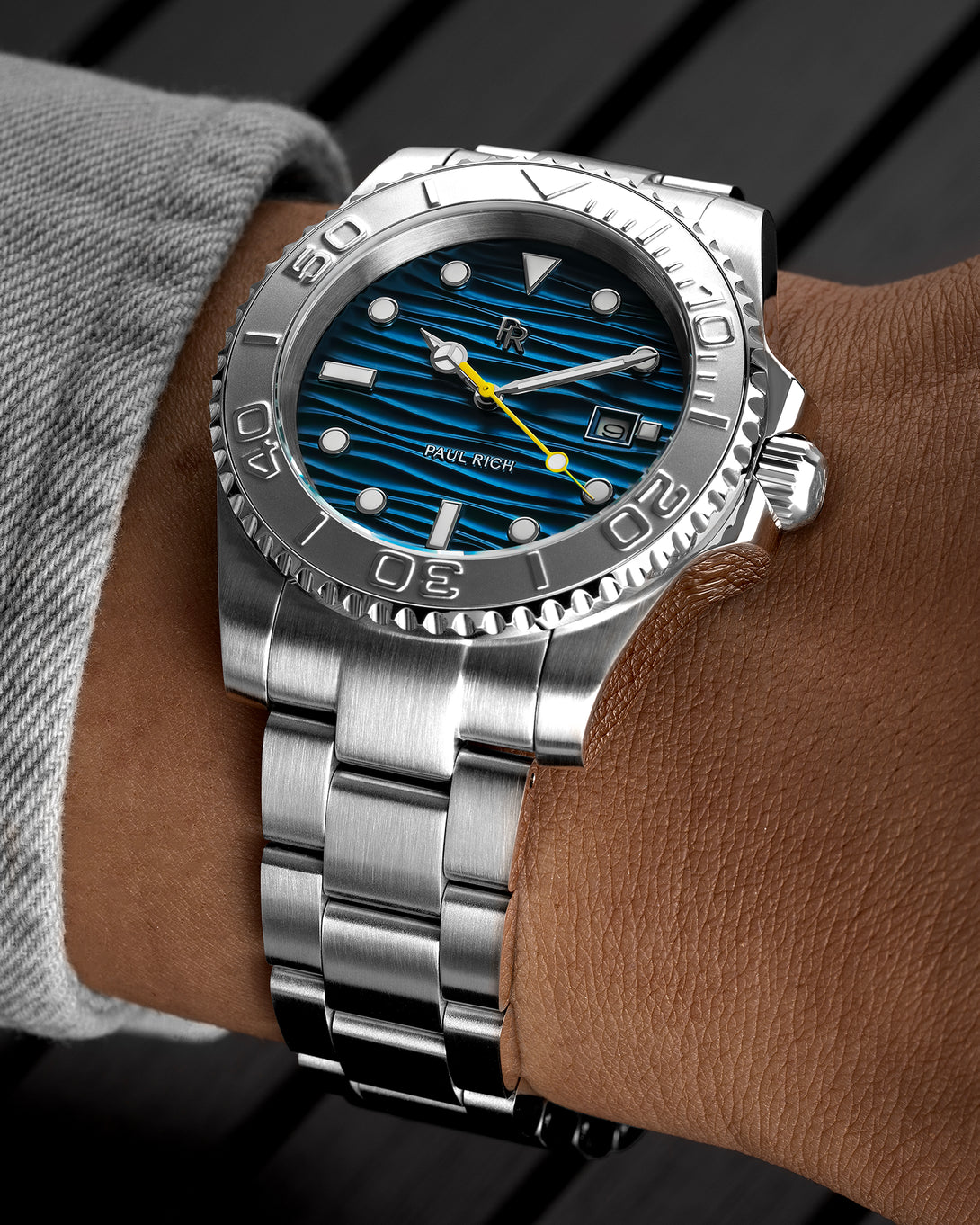 Stainless steel watch with a blue wave-textured dial and yellow seconds hand shown on a wrist, highlighting the bezel, markers, and brushed bracelet.