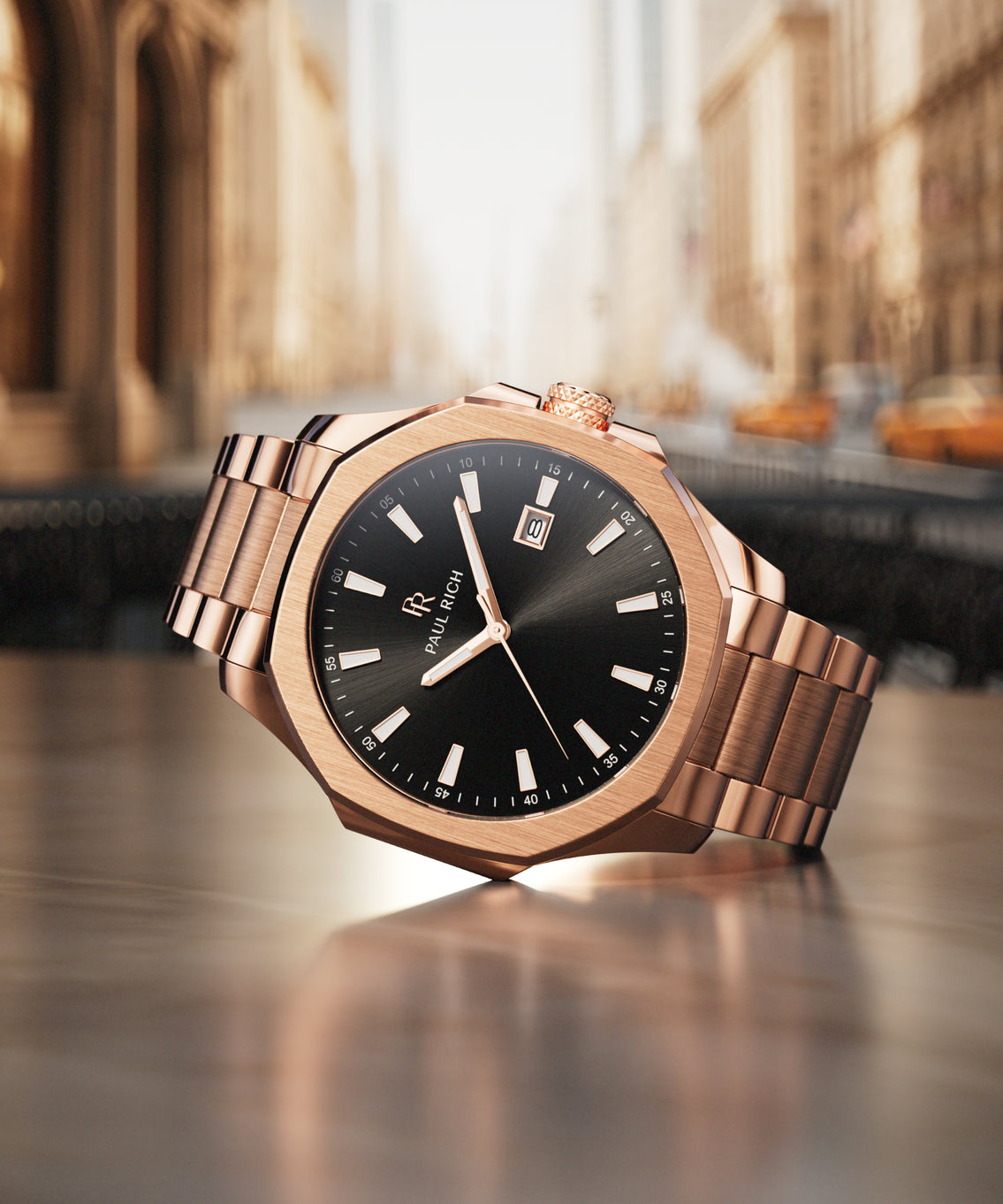 Rose gold watch with black dial on metal bracelet, displayed on a city street backdrop with blurred buildings and yellow taxis.