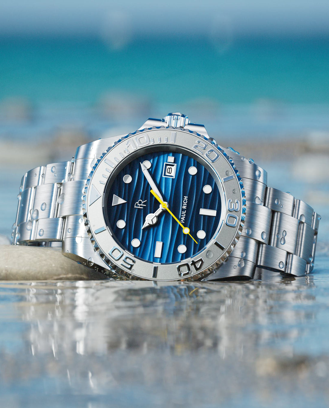 Silver dive watch with blue wave-textured dial, steel bracelet, rotating bezel, luminous markers, photographed in shallow water with ocean backdrop.