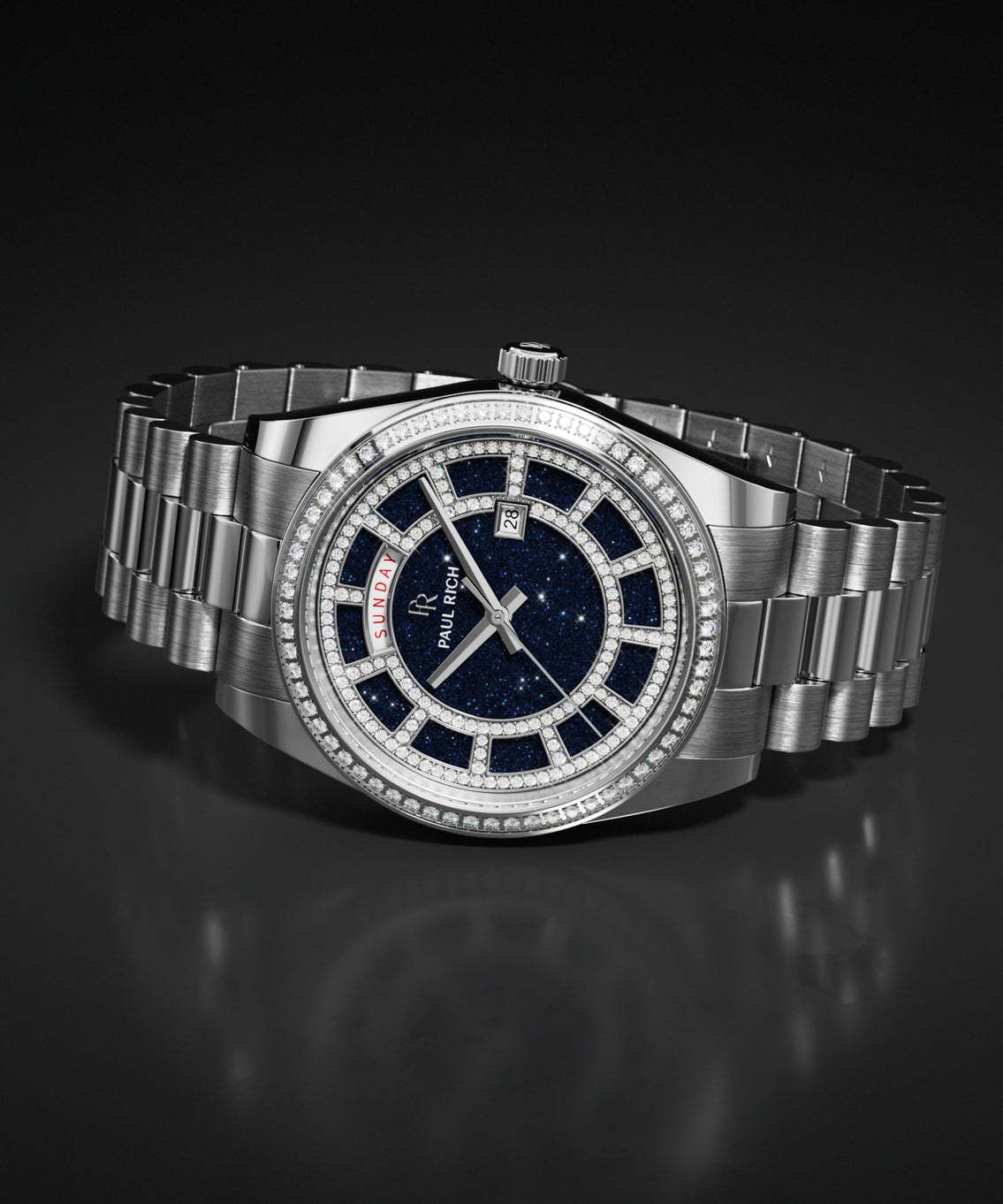 Blue aventurine dial watch with a silver metal bracelet, pavé-set bezel, day and date windows, and a polished case resting on a dark reflective surface.