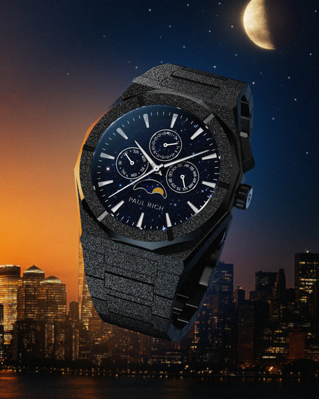 Frosted black stainless steel watch with blue aventurine dial, moonphase, and subdials, set against a city skyline at dusk with a moonlit sky.