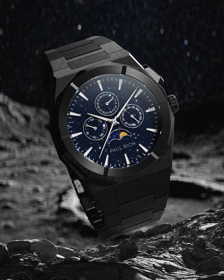 Brushed black stainless steel watch with blue aventurine dial, moonphase, and subdials, set against a rocky lunar surface and star-filled sky.