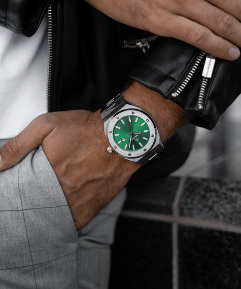Paul Rich Emperor's Emerald stainless steel watch with emerald green sunburst dial and brushed silver bezel, worn on wrist with a black leather jacket and light grey plaid trousers in an urban lifestyle setting.