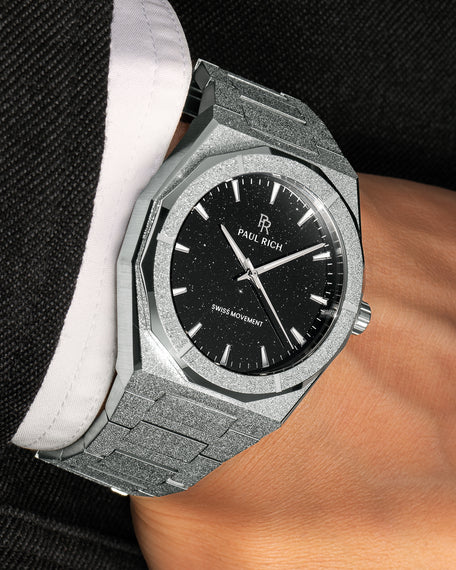 Silver watch with shimmering frosted case and black aventurine dial, worn with a white cuff and textured black jacket.