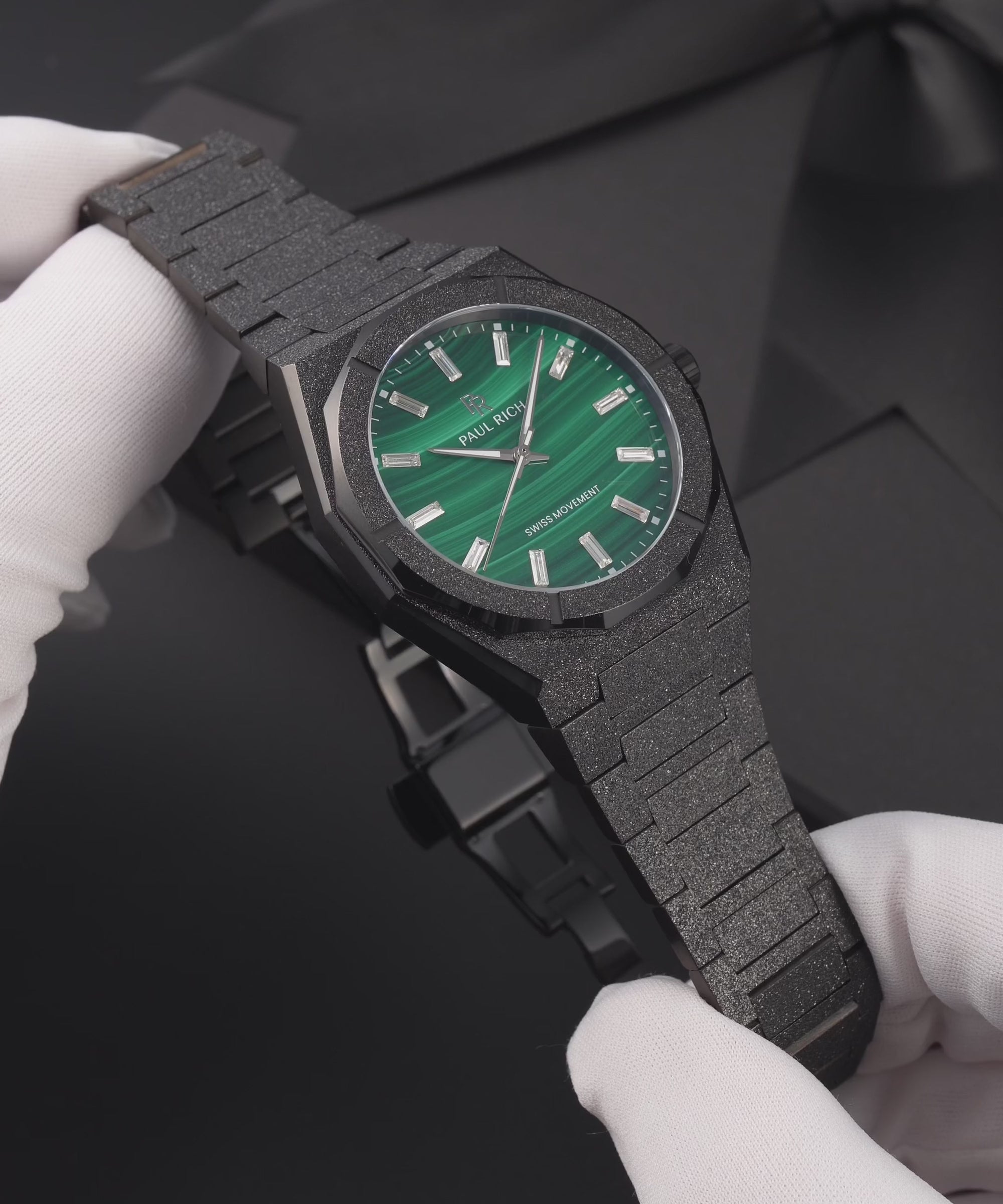 Black frosted Malachite Aurora watch with a green malachite dial, held in white gloves, worn on a wrist, standing on a black gift box and shown in close-up