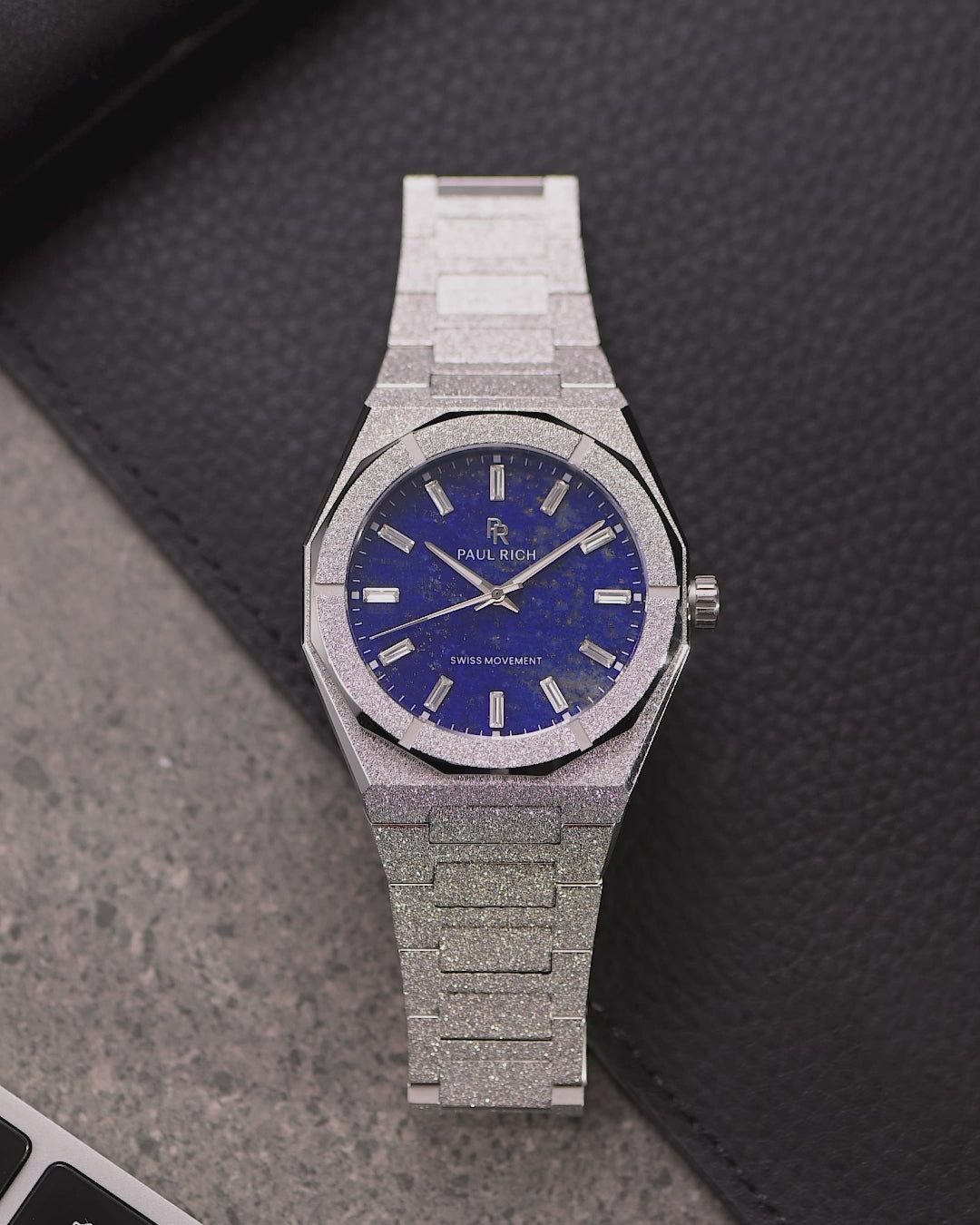 Silver frosted watch with a vivid blue dial lying beside a laptop
