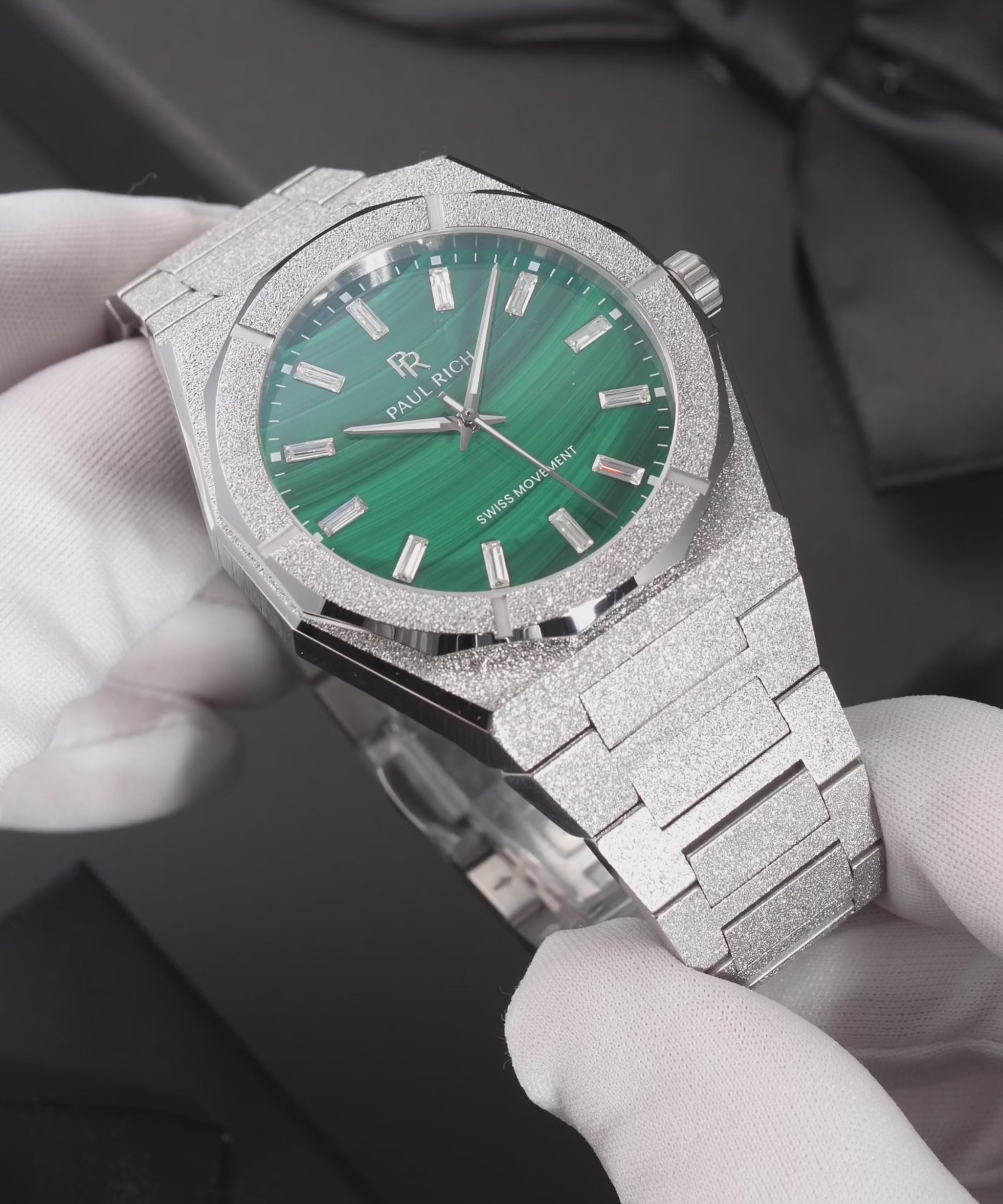 Silver frosted Malachite Aurora watch with a green malachite dial, held in white gloves, worn on a wrist, standing on a black gift box and shown in close-up