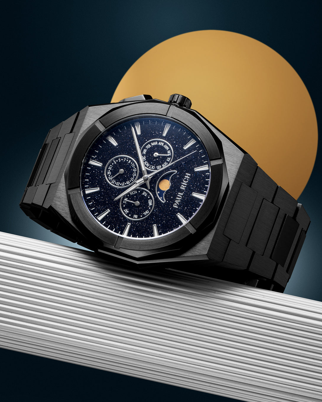 Black stainless steel watch with blue aventurine dial, moonphase, and subdials, displayed on a ribbed silver surface against a gold circle backdrop.