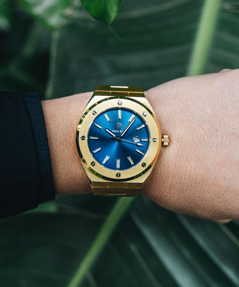 Paul Rich Royal Touch gold-tone watch with metallic blue sunburst dial and brushed bezel, worn on wrist in a tropical outdoor setting with lush green leaves in the background.